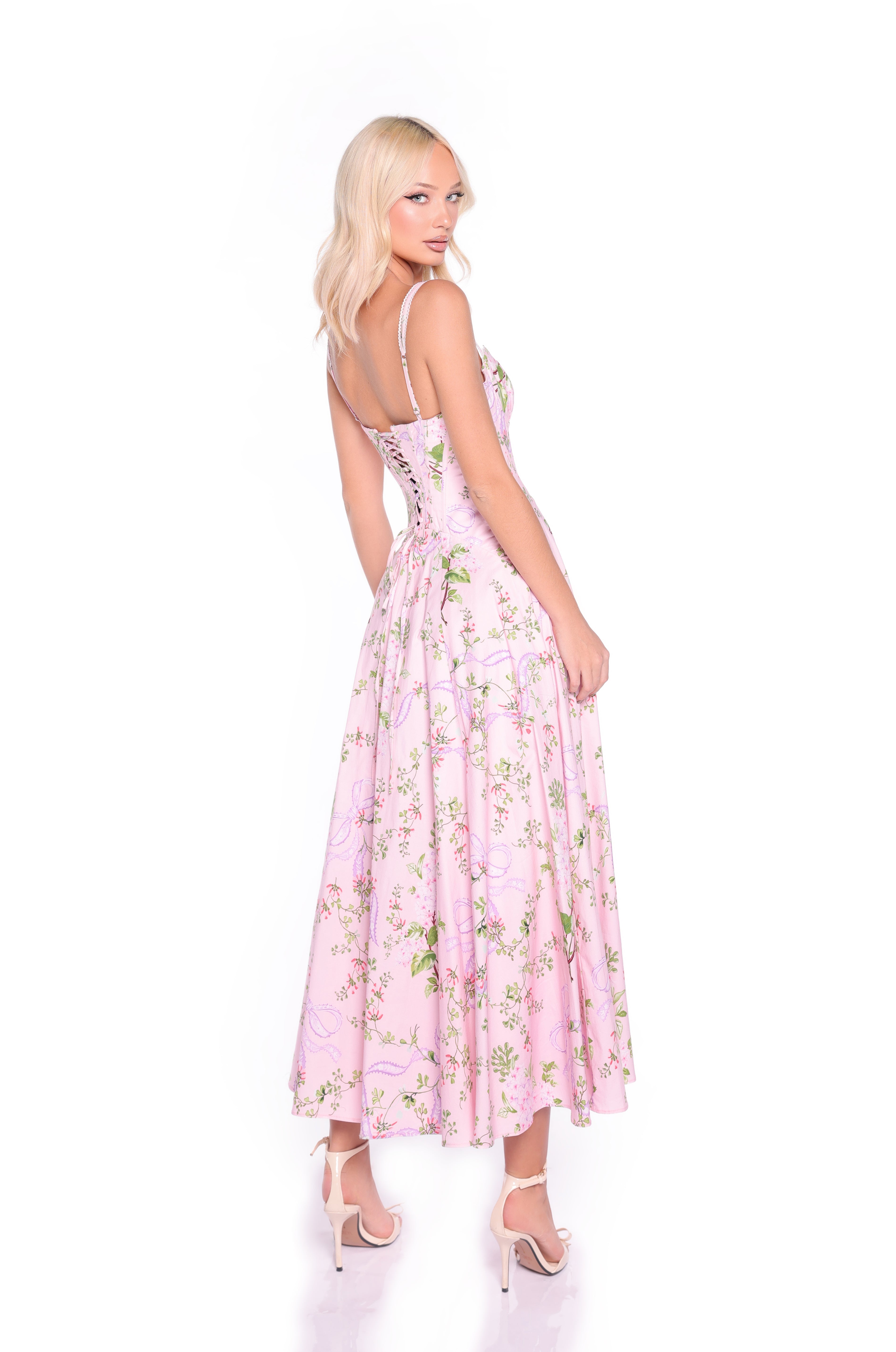 A woman with blonde hair models the Rosella Blush Pink Floral & Bow Print Casual Dress and white heels, posing with her back turned and looking over her shoulder against a white background.